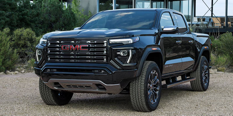 Front three-quarter view of a black GMC pickup truck parked outdoors on a gravel surface, showcasing a bold grille, chrome accents, and off-road tires in a residential setting.