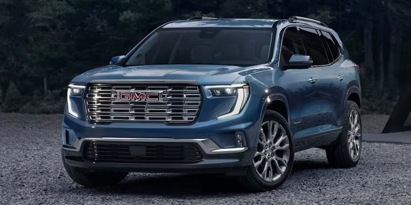 Front three-quarter view of a used blue GMC SUV parked outdoors on a gravel surface, featuring a bold chrome grille, LED headlights, and alloy wheels.
