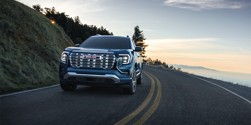Front view of a blue GMC Terrain driving on a winding coastal road at sunset, with a hillside on one side and an ocean view in the background, highlighting its bold grille and LED lighting.