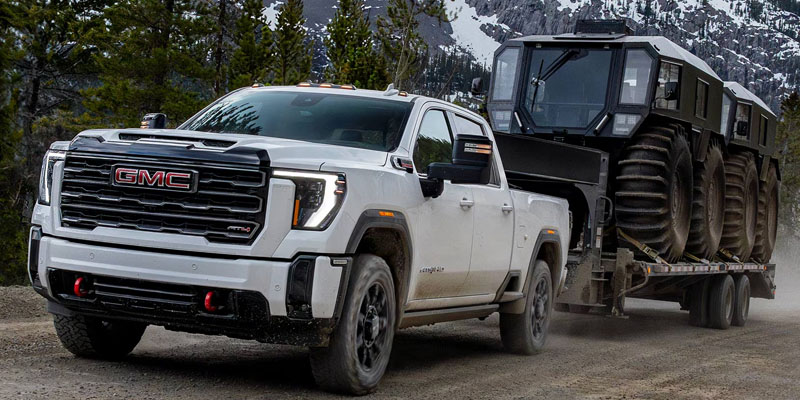 White GMC heavy-duty pickup truck towing large off-road equipment trailer through a mountain landscape.